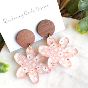 May include: A pair of pink floral earrings with a wooden accent. The earrings are shaped like a flower with five petals and have a small wooden circle at the top.