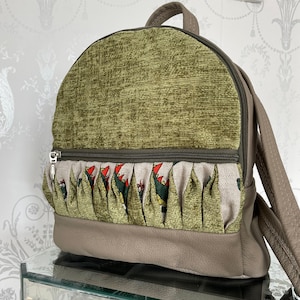 May include: A small backpack with a rounded top, featuring a textured olive green fabric on the top half and a light brown leather-like material on the bottom. The front has a pleated section with a repeating pattern of green and red figures.