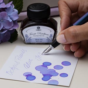 May include: A bottle of "Blue Iris" fountain pen ink next to a pen writing on a white paper. The ink is a vibrant blue-purple color, with the words "Octopus Fluids" and "Blue Iris" written on the paper. A bouquet of blue-purple flowers is in the background.