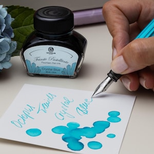 Könnte beinhalten: Eine Flasche "Fraise Pastelintte" Crystal Blue Füllertinte neben einem türkisfarbenen Füllfederhalter, der auf weißes Papier schreibt. Die Tinte hat eine leuchtende türkisfarbene Farbe. Das Papier hat Tintenkleckse und die Worte "Octopus Fluids Crystal Blue".