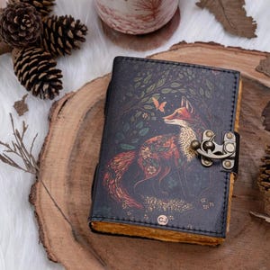 Peut inclure: Un journal en cuir foncé avec une illustration de renard et de papillon. Le journal est orné d'un fermoir en métal et de pages jaunies. La couverture présente une scène de forêt détaillée. Il est posé sur une surface en bois.