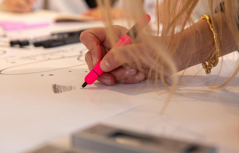 Puede incluir: Una mano sosteniendo un rotulador rosa y dibujando sobre una superficie blanca. La persona lleva una pulsera dorada en la mu&ntilde;eca.