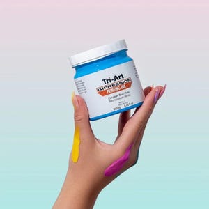 May include: A jar of Tri-Art Impression Printing Ink in Cerulean Blue. The jar is held in a hand with streaks of yellow and purple paint. The jar has a white lid and a blue base, with the text "Tri-Art Impression Printing Ink" on the label. The jar contains 8.45 fl oz.