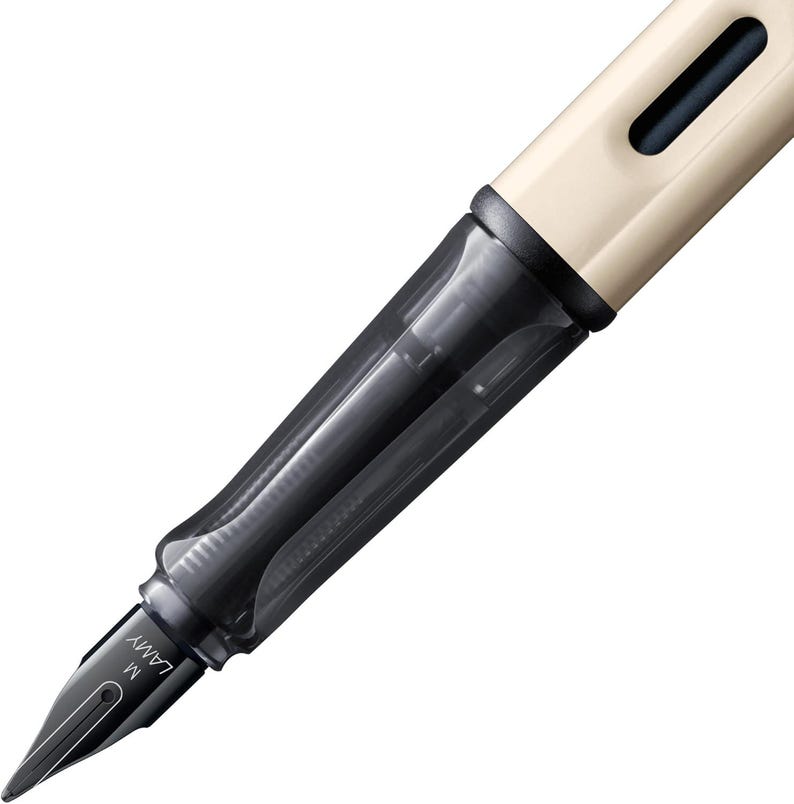 Puede incluir: Una pluma estilogr&aacute;fica negra y blanca con un cartucho de tinta transparente y una tapa beige. La pluma tiene una punta de metal con la etiqueta "LAMY M".