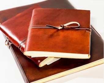 May include: Three leather-bound journals with a brown leather cover and a brown leather strap with a knot. The journals are stacked on top of each other.