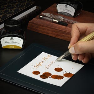 May include: A close-up of a person writing with a gold fountain pen on a white paper. The paper has brown ink splatters and the words "Octopus Fluids Bronze". Two ink bottles and a glass pen are also visible. The ink bottles have black caps and white labels.