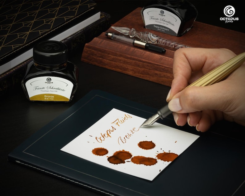 May include: A close-up of a person writing with a gold fountain pen on a white paper. The paper has brown ink splatters and the words "Octopus Fluids Bronze". Two ink bottles and a glass pen are also visible. The ink bottles have black caps and white labels.