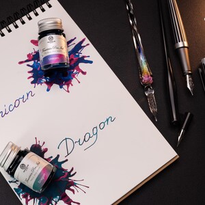 Pu&ograve; includere: Due piccoli flaconi di vetro di inchiostro per penna stilografica, uno etichettato "Unicorno" e l'altro "Drago". I flaconi sono su un taccuino bianco con rilegatura a spirale nera. Il taccuino &egrave; su una superficie nera con una variet&agrave; di strumenti di scrittura, tra cui una penna stilografica di vetro, una penna stilografica nera e una penna stilografica nera e argentata.