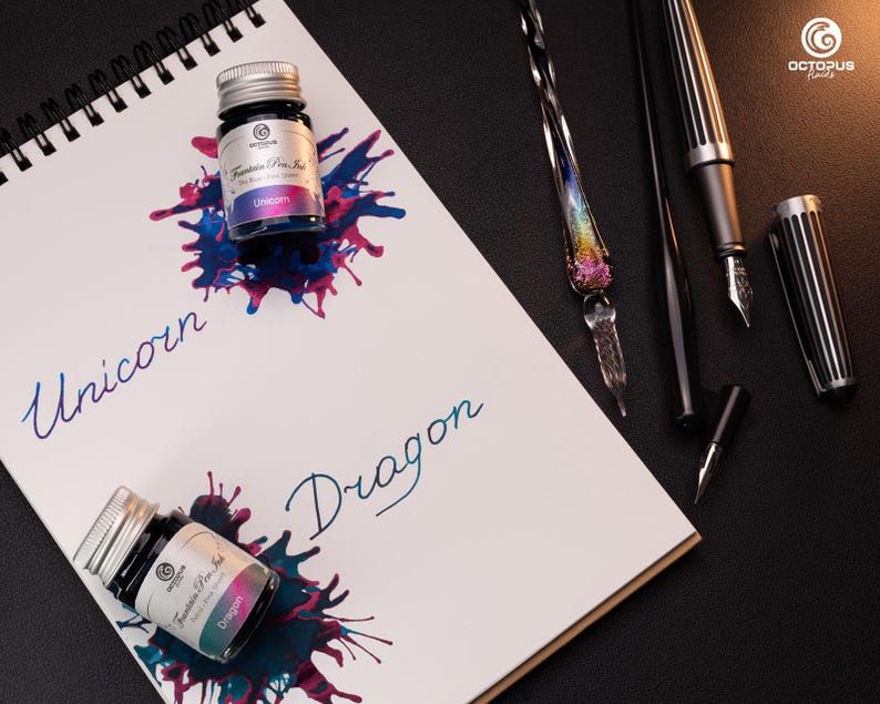 Pu&ograve; includere: Due piccoli flaconi di vetro di inchiostro per penna stilografica, uno etichettato "Unicorno" e l'altro "Drago". I flaconi sono su un taccuino bianco con rilegatura a spirale nera. Il taccuino &egrave; su una superficie nera con una variet&agrave; di strumenti di scrittura, tra cui una penna stilografica di vetro, una penna stilografica nera e una penna stilografica nera e argentata.