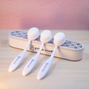May include: Three white oval makeup brushes with soft, white bristles are displayed in a gray and white holder. The brushes have long, white handles with the text "prim studio" printed on them. The holder has a geometric design.