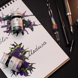 Pu&ograve; includere: Due bottiglie di inchiostro per penna stilografica, una etichettata "Goblin" e l'altra "Medusa". Le bottiglie sono su un taccuino bianco con schizzi di inchiostro nero. Ci sono anche tre strumenti di scrittura: una penna stilografica nera, una penna a immersione nera e una penna a immersione di vetro con vetro color arcobaleno.