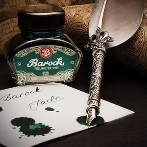 Op de afbeelding: Een inktfles in vintage-stijl met een zwarte dop en een groen en wit etiket met de tekst "Barock Follhertinte Jade". Een decoratieve zilveren pen met een veer en de woorden "Barock Jade" geschreven op wit papier met inktvlekken.