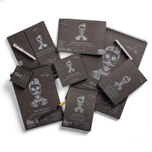May include: A collection of black sketchbooks and pads with a skull illustration. The sketchbooks are in various sizes, including spiral-bound and square formats. The text on the covers reads "Black sketch pad" and "Sketch #authenticbook". Pens are also included.