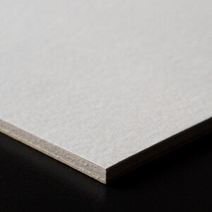 May include: Close-up of a white cardstock sheet with a textured surface. The edge of the sheet is visible, showing a slightly darker gray layer.