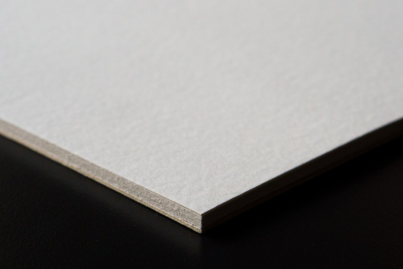 May include: Close-up of a white cardstock sheet with a textured surface. The edge of the sheet is visible, showing a slightly darker gray layer.