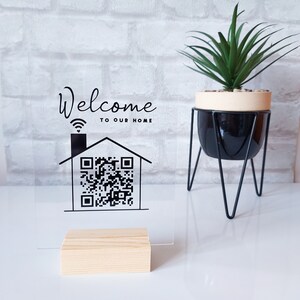Könnte beinhalten: Transparente Acryltafel mit der Aufschrift "Welcome to our home" und einem QR-Code in einem Hausumriss. Sie steht auf einem Holzsockel. Rechts befindet sich ein schwarz-weißer Pflanztopf mit einer grünen Pflanze.