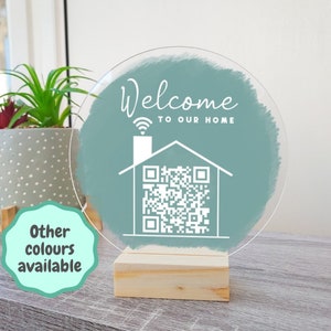Peut inclure: Panneau rond en acrylique transparent avec l'inscription "Welcome to our Home" et un motif de maison avec un code QR. Le panneau est monté sur une base en bois naturel. Un coup de pinceau de couleur bleu-vert est derrière le texte et le motif.