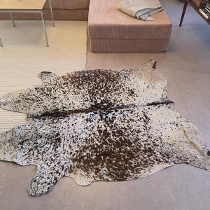 May include: A cowhide rug featuring an irregular, organic shape with a speckled pattern of dark brown and black spots distributed across a white background. The hide displays a natural, asymmetrical silhouette with a distinct dark central spine line.