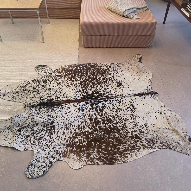 May include: A cowhide rug featuring an irregular, organic shape with a speckled pattern of dark brown and black spots distributed across a white background. The hide displays a natural, asymmetrical silhouette with a distinct dark central spine line.