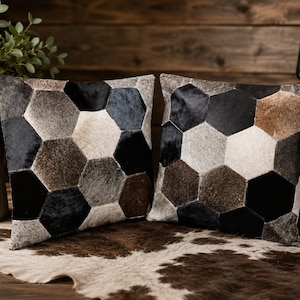 Könnte beinhalten: Zwei quadratische Zierkissen mit einem geometrischen Muster aus vernähten sechseckigen Flicken. Die Flicken bestehen aus Fell in verschiedenen Schwarz-, Weiß-, Grau- und Brauntönen und bilden eine strukturierte, mehrfarbige Mosaikoberfläche.
