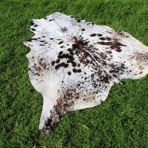 May include: A natural cowhide rug featuring an irregular, organic shape with a white base color and a dense pattern of dark brown and black spots distributed across the surface, showcasing the unique texture and markings of the animal hide.