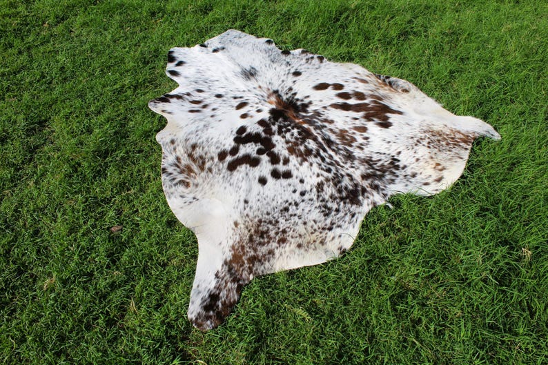 May include: A natural cowhide rug featuring an irregular, organic shape with a white base color and a dense pattern of dark brown and black spots distributed across the surface, showcasing the unique texture and markings of the animal hide.