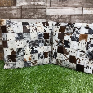 May include: Two square throw pillows with a patchwork design made from cowhide. The pillows are brown, white, and black with a checkered pattern.