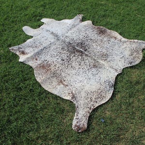 May include: A natural cowhide rug featuring an irregular, organic shape with a speckled pattern of white, grey, and dark brown hair. The hide displays a dense distribution of small, dark spots across the surface, creating a mottled appearance.