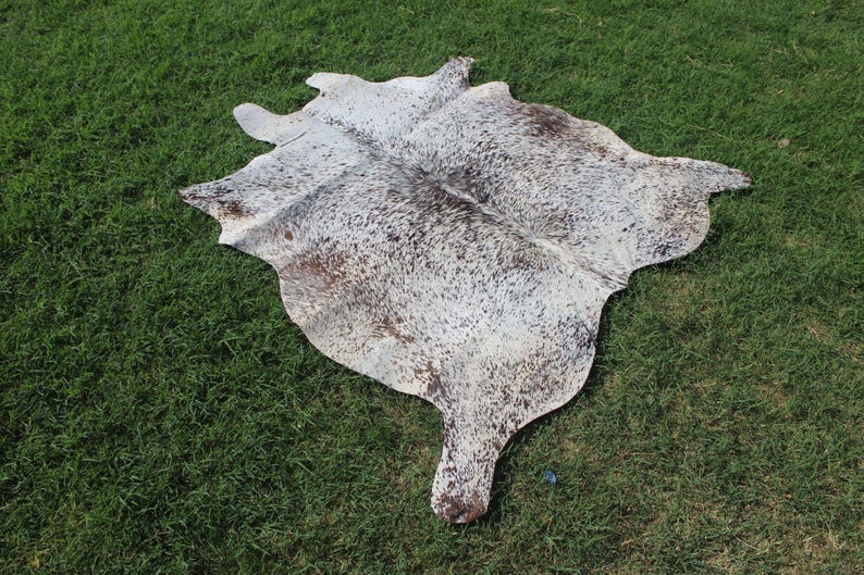 May include: A natural cowhide rug featuring an irregular, organic shape with a speckled pattern of white, grey, and dark brown hair. The hide displays a dense distribution of small, dark spots across the surface, creating a mottled appearance.