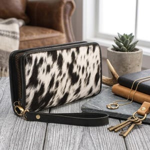 May include: A rectangular wallet featuring a black and white cowhide pattern with a textured hair-on-hide finish. The wallet includes a black leather wrist strap attached to a gold-toned metal zip pull and a dark brown leather trim along the edges.