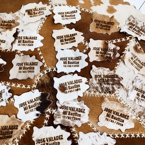 May include: A collection of 18 cowhide place cards with the text "JOSE VALADEZ Mi Bautizo 12/02/2023" engraved on each. The place cards are arranged on a brown and white cowhide surface.