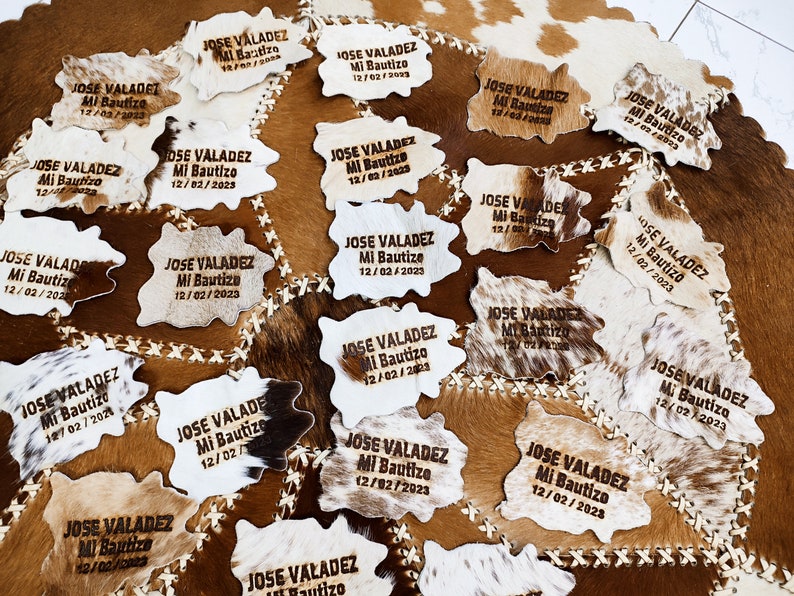 May include: A collection of 18 cowhide place cards with the text "JOSE VALADEZ Mi Bautizo 12/02/2023" engraved on each. The place cards are arranged on a brown and white cowhide surface.