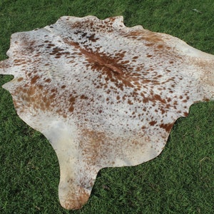May include: A brown and white cowhide rug with a spotted pattern. The rug is laid out on a green grassy surface.