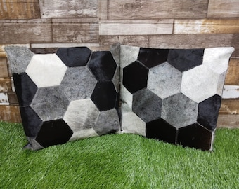 May include: Two square throw pillows with a geometric pattern made of black, white, and gray cowhide. The pillows are arranged side by side on a green grassy surface.