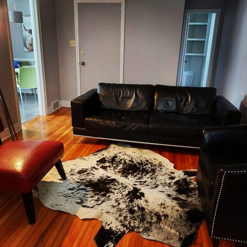 May include: A black leather sofa with thin metal legs, a matching black leather armchair with silver nailhead trim, a red upholstered chair, and a cowhide rug featuring a black and white spotted pattern on a hardwood floor.