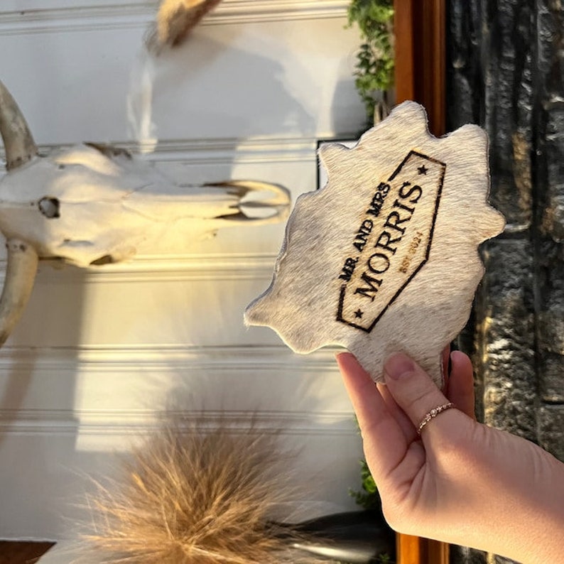 May include: A white cowhide coaster with a brown, rectangular, engraved label that reads "Mr. and Mrs. Morris Est. 2021".