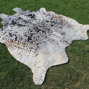 May include: A natural cowhide rug featuring a salt and pepper pattern with a mix of white, grey, black, and light brown spots. The hide has an irregular, organic shape with a smooth texture and a distinct mottled appearance across the entire surface.