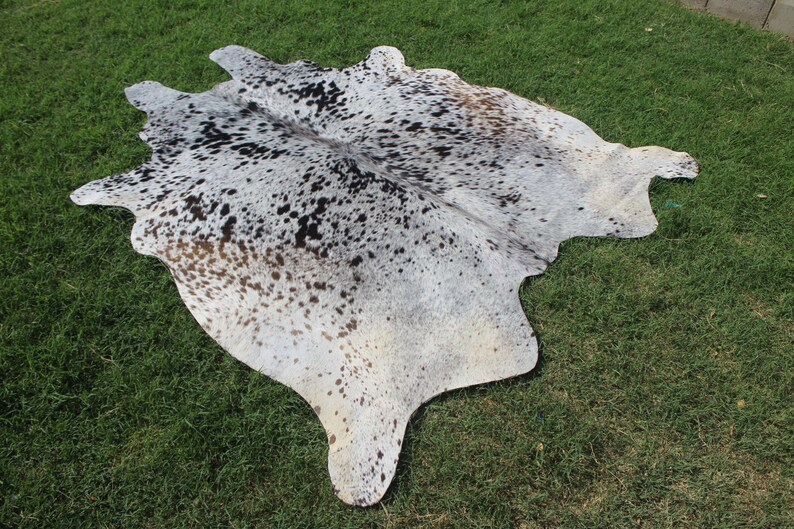 May include: A natural cowhide rug featuring a salt and pepper pattern with a mix of white, grey, black, and light brown spots. The hide has an irregular, organic shape with a smooth texture and a distinct mottled appearance across the entire surface.
