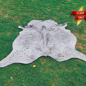 May include: A cowhide rug featuring a natural, irregular shape with a white base color and scattered grey and black speckles throughout the surface. A red banner with the white text FLASH SALE is positioned in the upper right corner of the image.