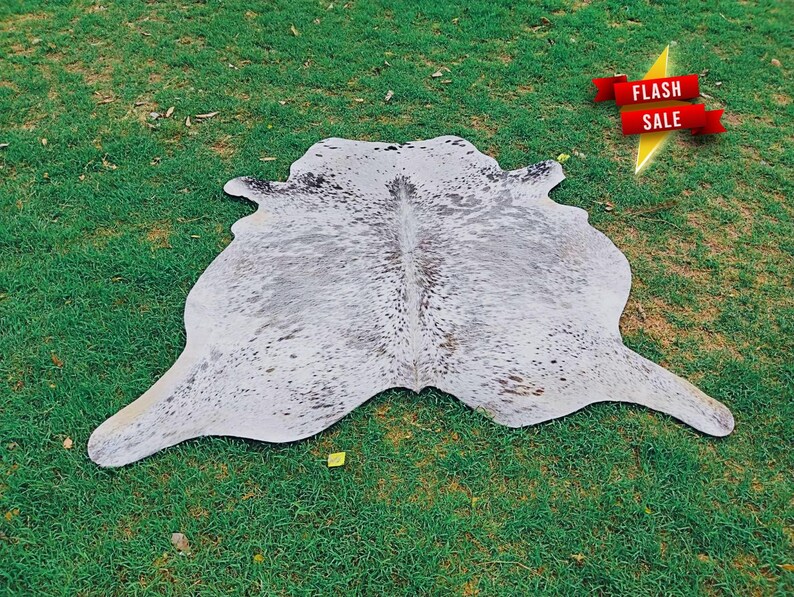May include: A cowhide rug featuring a natural, irregular shape with a white base color and scattered grey and black speckles throughout the surface. A red banner with the white text FLASH SALE is positioned in the upper right corner of the image.
