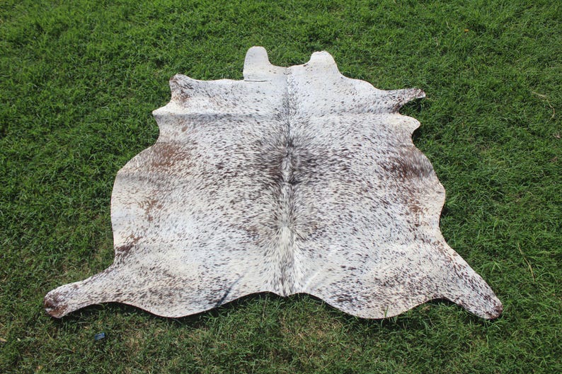 May include: A natural cowhide rug featuring an irregular, organic shape with a speckled pattern of white, grey, and dark brown spots. The hide displays a dense, short-haired texture across its entire surface, showcasing a unique, mottled color variation.