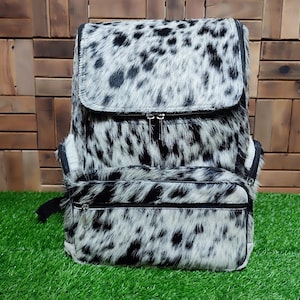 May include: A black and white cowhide backpack with a zippered front pocket. The backpack has a unique spotted pattern.
