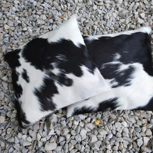 May include: Two black and white cowhide throw pillows with a soft, fuzzy texture. The pillows are on a bed of small gray rocks.