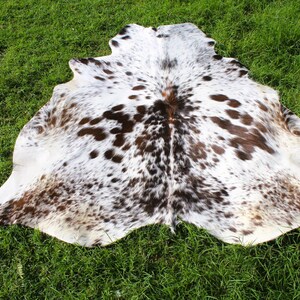 May include: A cowhide rug featuring a natural pattern of white, brown, and black spots. The hide has an irregular, organic shape with a smooth surface texture, displaying a mix of dark brown and black patches scattered across a white base.