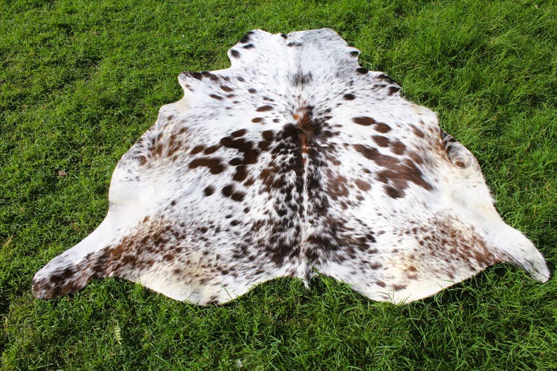 May include: A cowhide rug featuring a natural pattern of white, brown, and black spots. The hide has an irregular, organic shape with a smooth surface texture, displaying a mix of dark brown and black patches scattered across a white base.
