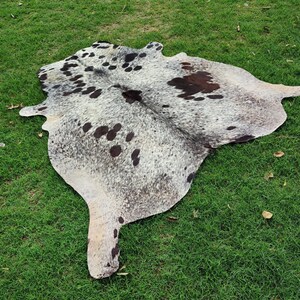 May include: A natural cowhide rug featuring a mottled pattern of white, grey, and black speckles with larger dark brown and black patches distributed across the irregular, organic shape of the hide, showcasing a smooth, flat, and textured surface.