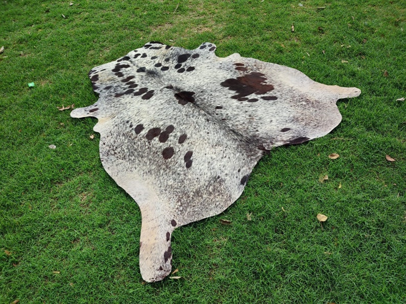 May include: A natural cowhide rug featuring a mottled pattern of white, grey, and black speckles with larger dark brown and black patches distributed across the irregular, organic shape of the hide, showcasing a smooth, flat, and textured surface.