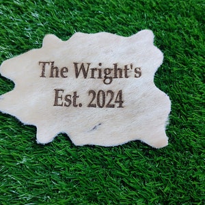 May include: A white leather patch with the text "The Wright's Est. 2024" burned into the surface. The patch is sitting on a green artificial grass surface.