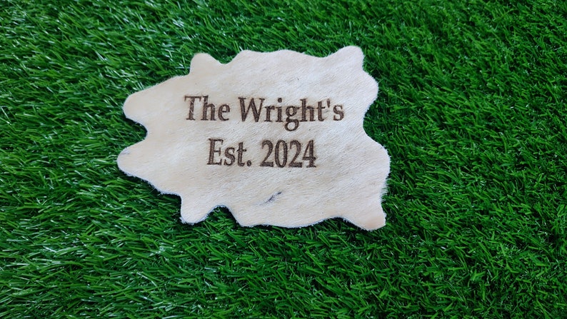 May include: A white leather patch with the text "The Wright's Est. 2024" burned into the surface. The patch is sitting on a green artificial grass surface.