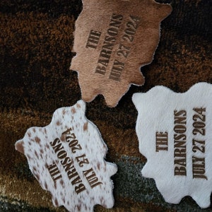 May include: Three cowhide coasters with the text "THE BARNSONS JULY 27 2024" engraved on each. The coasters are in different colors and patterns, including brown, white, and brown with white spots.